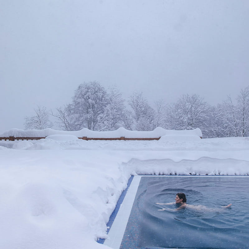 Location Winter Wellbeing woman swimming in an outdoor pool surrounded by high snow with snow covered forest in the background